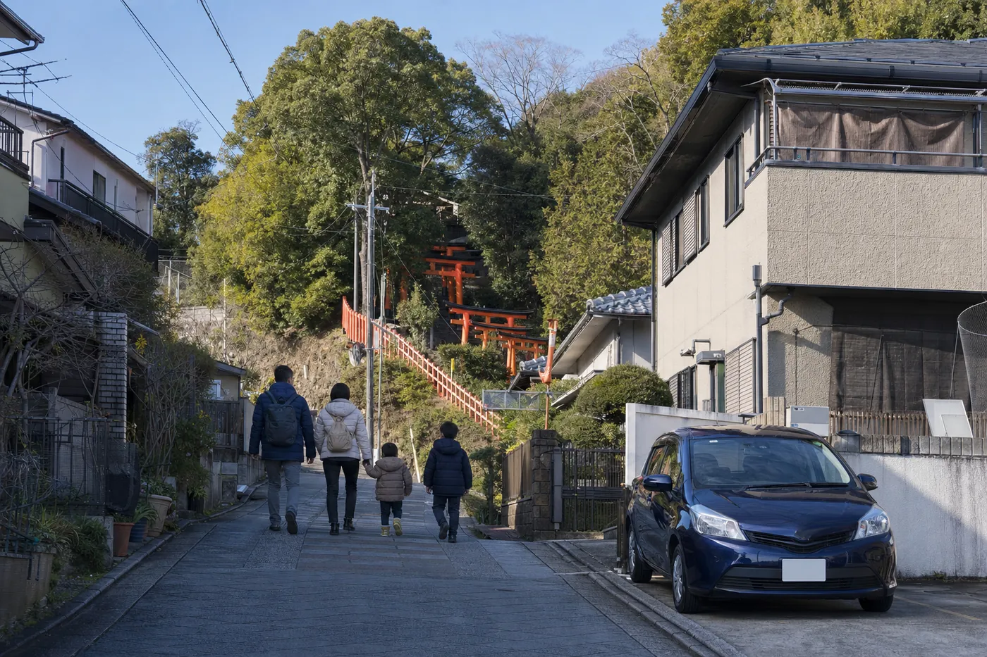 冬の晴れた朝、山のふもとの住宅街の坂道を家族4人が歩き、奥の木々の間に朱色の鳥居が連なって山を上っている。手前の住宅の駐車スペースには紺色の小型車が停まっている写真風のイメージ（AI生成）