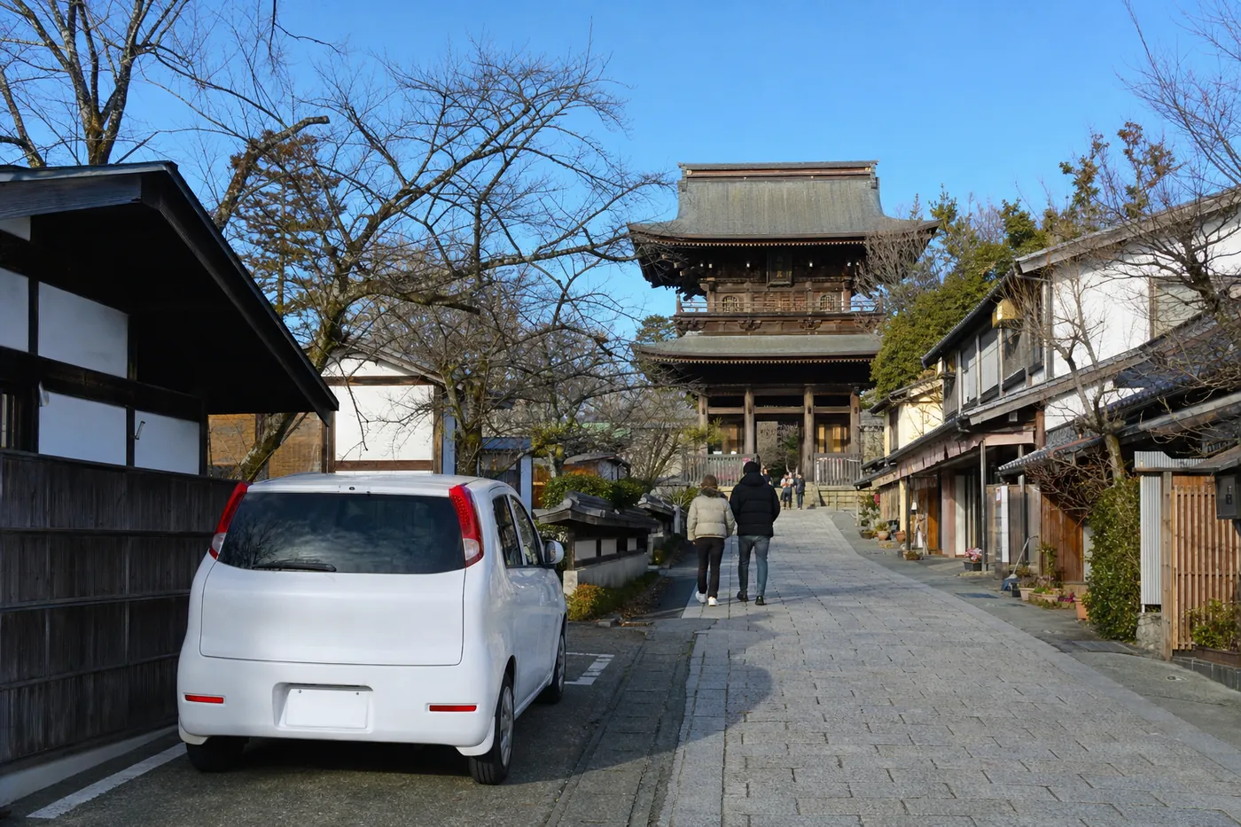 冬の晴れた朝、木造の店が並ぶ石畳の参道の先に大きな寺の山門が見え、コートを着た2人が歩いている。手前の駐車スペースには白い小型車が停まっている写真風のイメージ（AI生成）