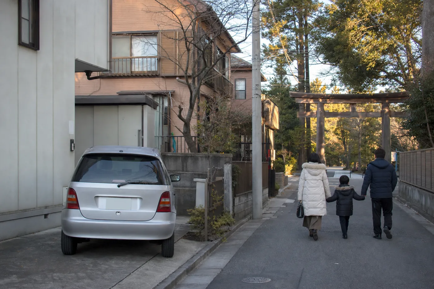 元日の朝、住宅の間の駐車スペースに銀色の車が停まり、冬のコートを着た家族3人が、常緑樹に囲まれた大きな木の鳥居に向かって歩いている写真風のイメージ（AI生成）