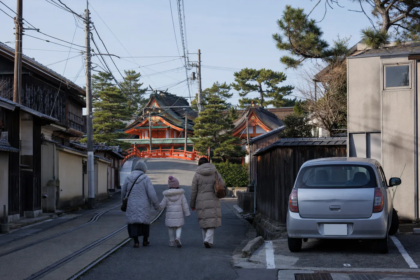元日の朝、路面電車の線路がある通りを、祖母・母・子の3人が、松の木の向こうに見える朱色の社殿と反り橋に向かって歩いている。右手前の駐車スペースには銀色の軽自動車が停まっている写真風のイメージ（AI生成）