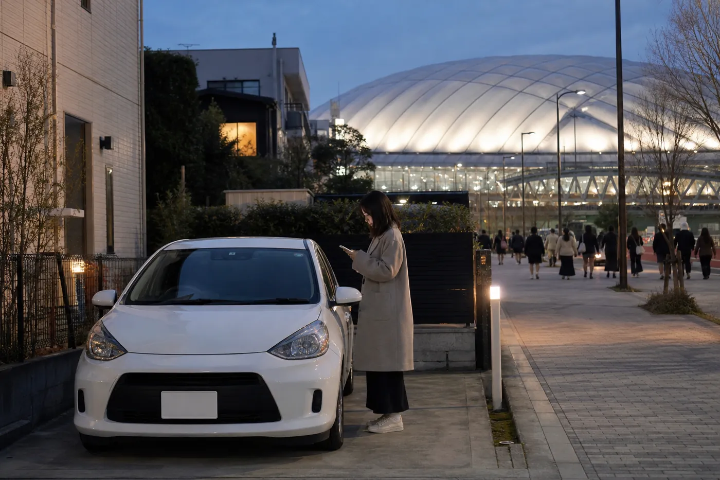 夕暮れの都心で、住宅の間の駐車スペースに白い車が停まり、女性がスマートフォンを見ている。奥では白いドームが光り、人が向かって歩いている写真風のイメージ（AI生成）