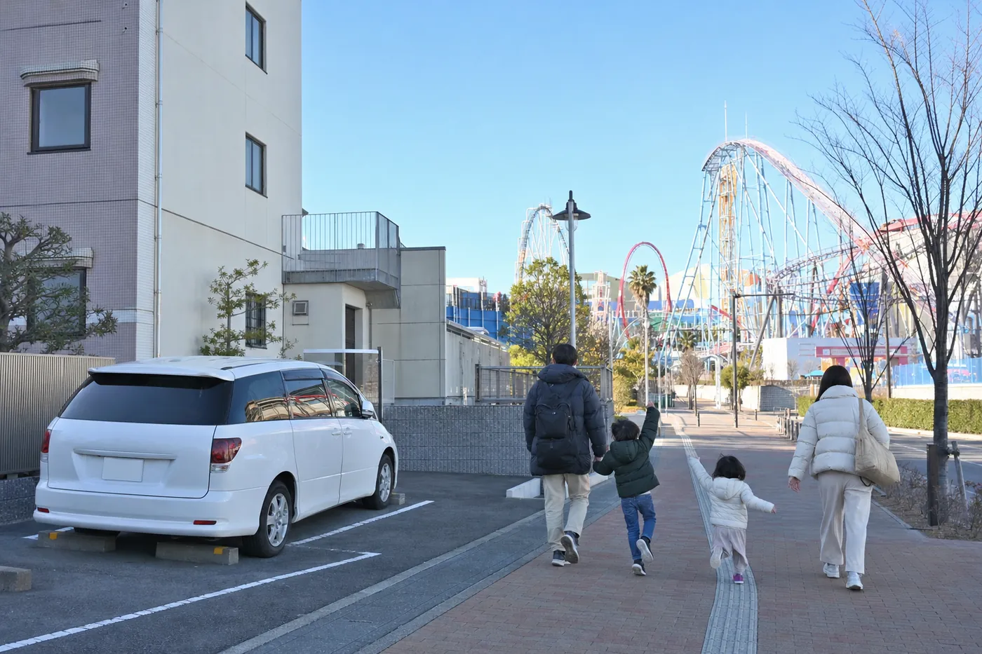 冬の晴れた朝、マンションの横の駐車場に白いミニバンが停まり、子どもが2人いる家族が、遠くにジェットコースターが見える遊園地に向かって広い歩道を歩いている写真風のイメージ（AI生成）