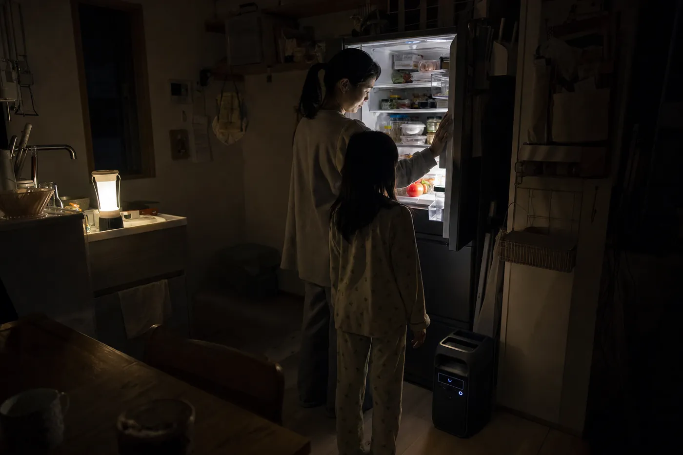 停電した夜のキッチンで、親子が明かりのついた冷蔵庫を開けている写真風のイメージ。冷蔵庫の横にポータブル電源が置いてある
