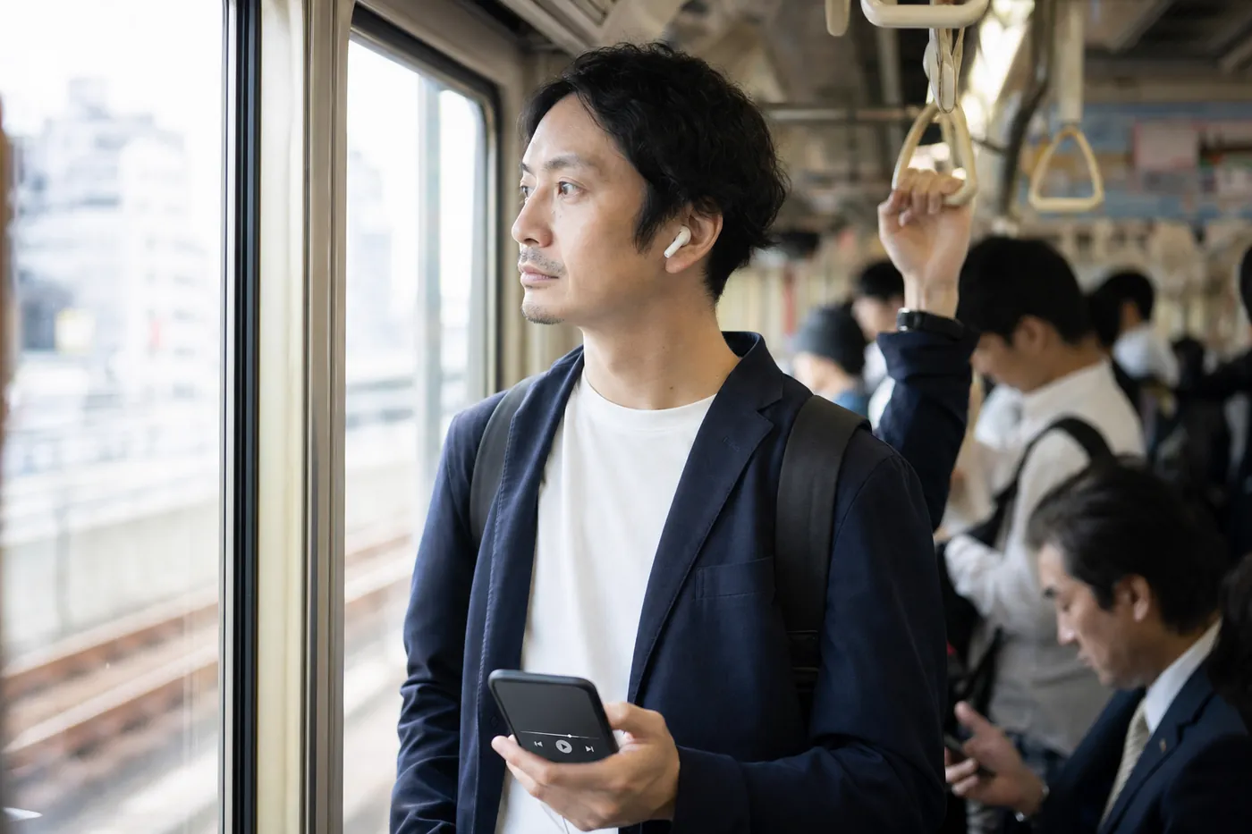 朝の通勤電車で、つり革につかまりワイヤレスイヤホンで何かを聴きながら窓の外を見ている人の写真風のイメージ(AI生成)