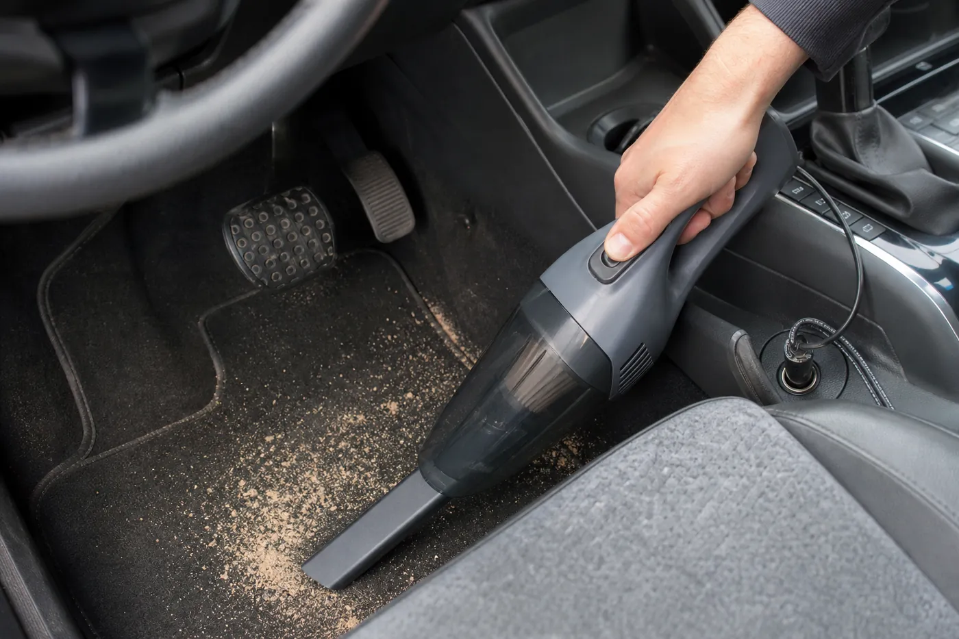 車のアクセサリーソケットにコードをつないだ小型のハンディクリーナーで、運転席の足元の砂を吸っている写真風のイメージ(AI生成)