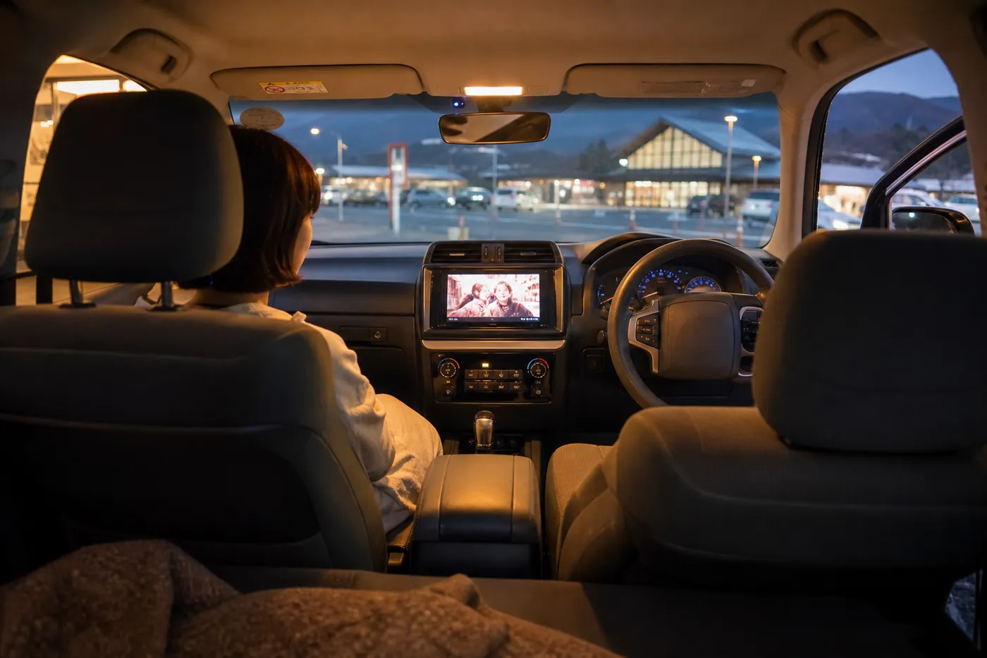 夕方の道の駅の駐車場に止めた車内で、助手席の人が純正ナビの画面で動画を見ているイメージ(AI生成)