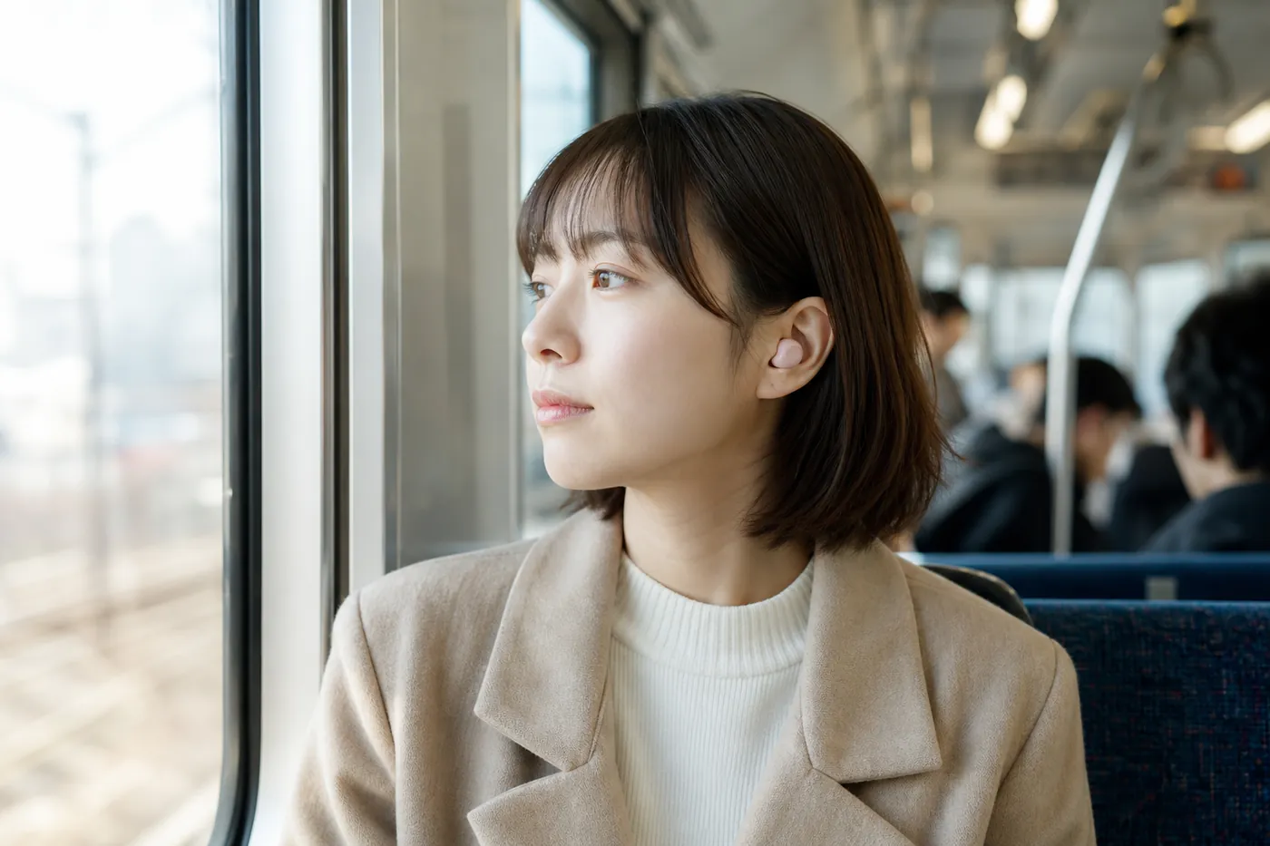朝の通勤電車で、小さなピンクのワイヤレスイヤホンを着けて窓の外を見る女性の写真風のイメージ