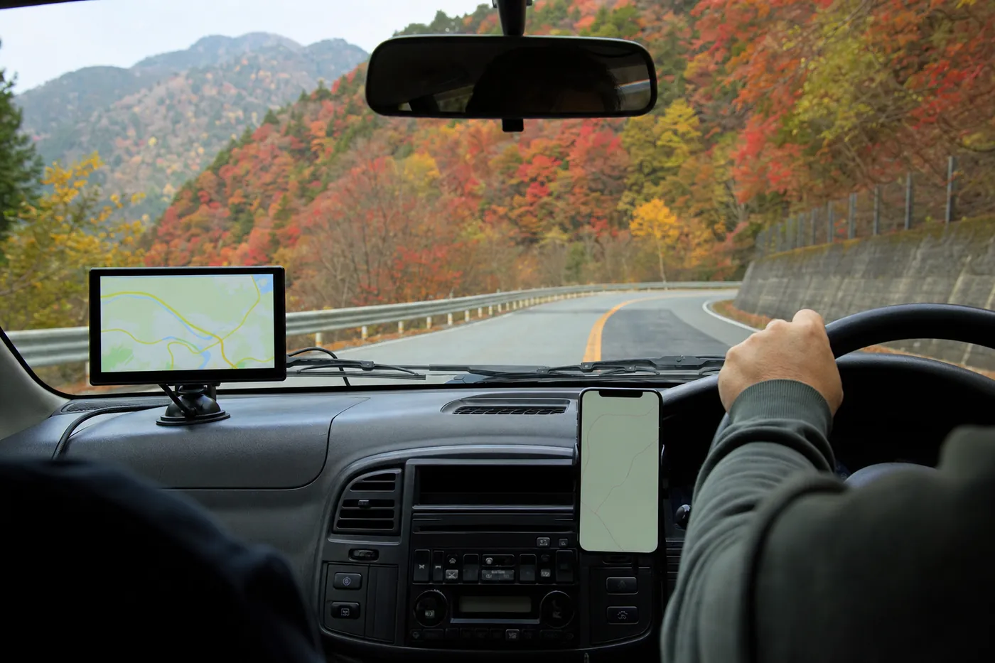 紅葉の山道を走る車内で、ダッシュボードのポータブルナビとスマホホルダーのスマホを並べた写真風のイメージ