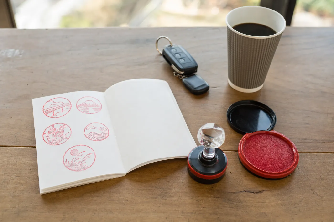 道の駅のテーブルに、スタンプを押した冊子とスタンプ台、車の鍵とコーヒーが置かれている写真風のイメージ（AI生成）
