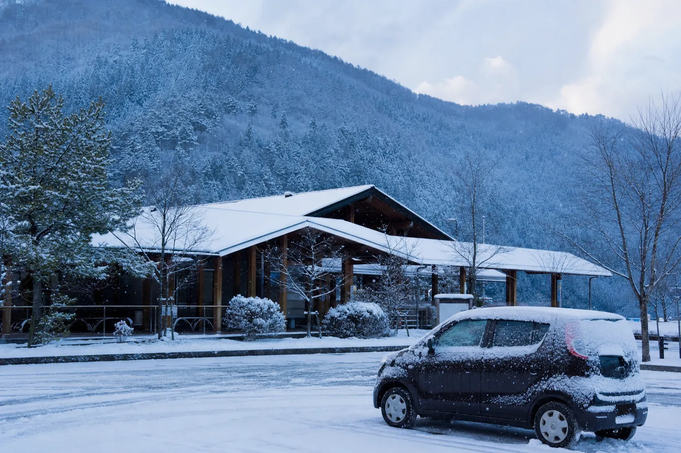 雪の積もった山あいの道の駅の駐車場に、雪をかぶった車が止まっている写真風のイメージ（AI生成）