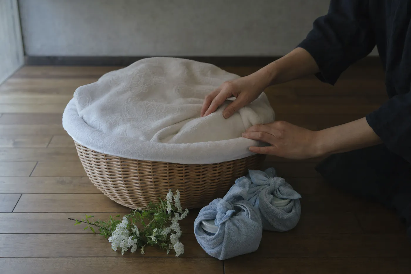 タオルを敷いたかごと、タオルで包んだ保冷剤、白い花を床に置き、かごの毛布を整える手元の写真風のイメージ