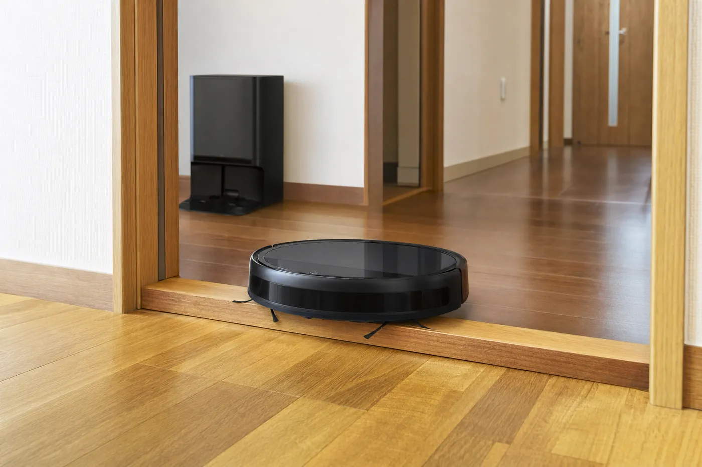 木の床の部屋と廊下の間の敷居を乗り越える黒いロボット掃除機と、奥に置かれた黒い全自動ドックのイメージ（AI生成）