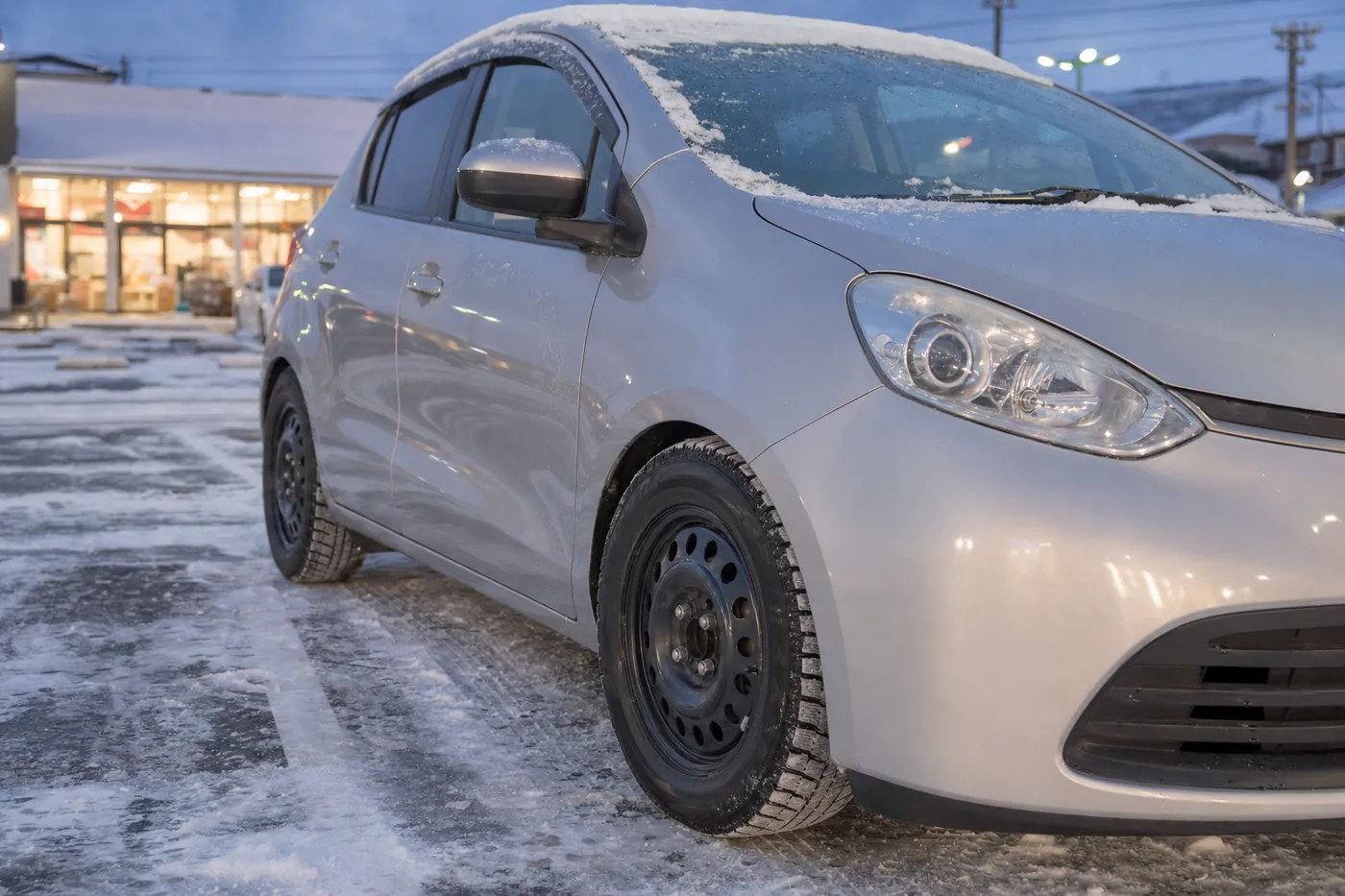 雪が積もったスーパーの駐車場に止めたシルバーのコンパクトカー。前輪にスタッドレスタイヤと黒いホイールが付いているイメージ(AI生成)