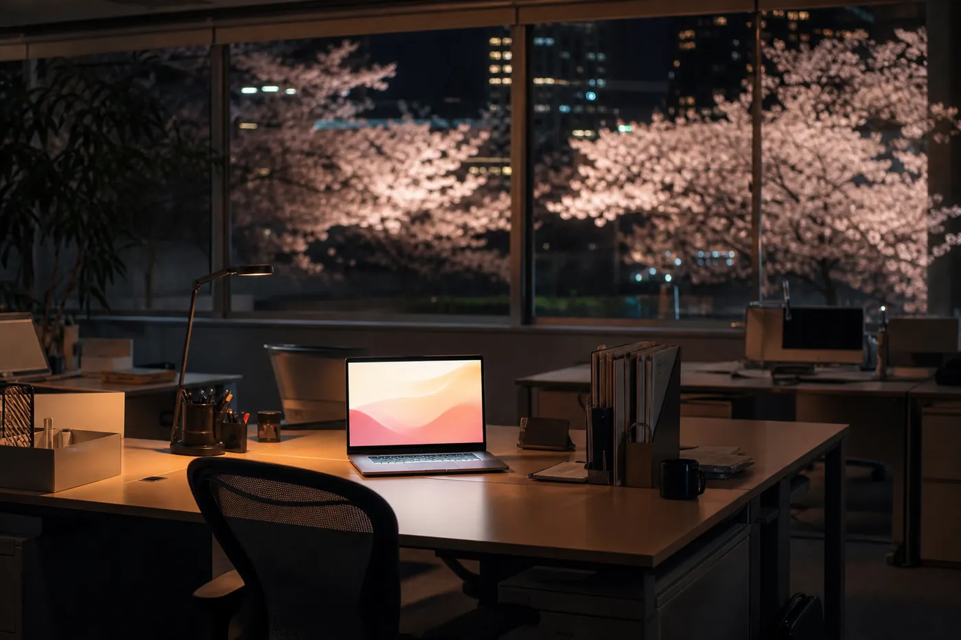 桜が見える夜のオフィスで、ノートパソコンだけが明るく光っている机の写真風のイメージ（AI生成）
